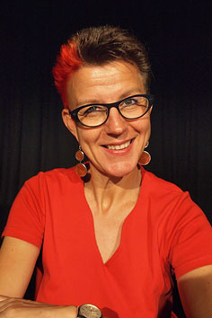 Foto von Andrea Haupt vor einem schwarzen Vorhang. Sie trägt ein rotes Top mit spitzem V-Ausschnitt und eine Brille. Ihre Haare sind kurz und zum Teil rot gefärbt. Sie lächelt in die Kamera. Sie trägt Ohrringe mit Metallscheiben.