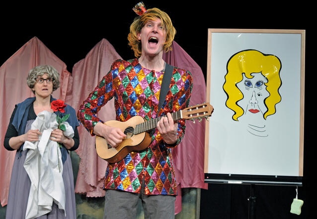 B&uuml;hnenfoto: Links neben einem Whiteboard, auf mit Magneten aus verschiedenen Versatzst&uuml;cken ein Gesicht zusammengeklebt ist, steht Dr. Tale als Prinz in einer bunten Glitzerjacke und Kr&ouml;nchen auf der Per&uuml;cke mit Gitalele und singt. Links schr&auml;g hinter ihm steht Mrs. Fairy, schon verkleidet als alte Fee mit Rose und wartet darauf, dass Dr. Tale fertig wird, damit die Szene losgehen kann.