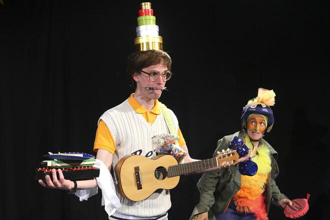 B&uuml;hnenfoto: In der Mitte steht Bert, auf seinem Kopf balanciert er einen Stapel runder Boxen und auf der linken Hand ein Glockenspiel, darauf eine Melodika und darauf einen Pappteller mit Krams. In der rechten Hand h&auml;lt er eine Teekanne und an einem Finger baumelt eine Tasse. Auf der Gitalele, die an ihm h&auml;ngt liegt eine T&uuml;te Konfetti. Zwischen den Lippen h&auml;lt er einen L&ouml;ffel und eine Gabel. Der Happy Bird steht rechts neben ihm und schaut ihn belustigt an.